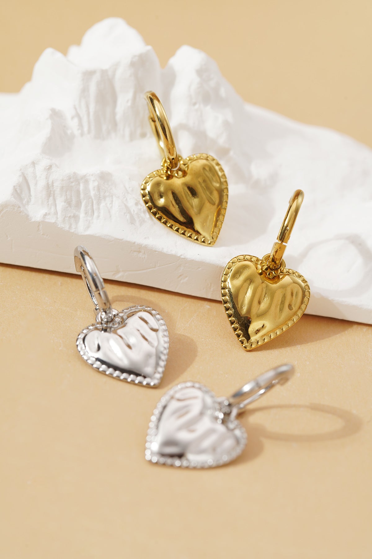 Gold and silver Sculpted Coquette heart charm hoop earrings with a beaded border, resting on a white textured base.