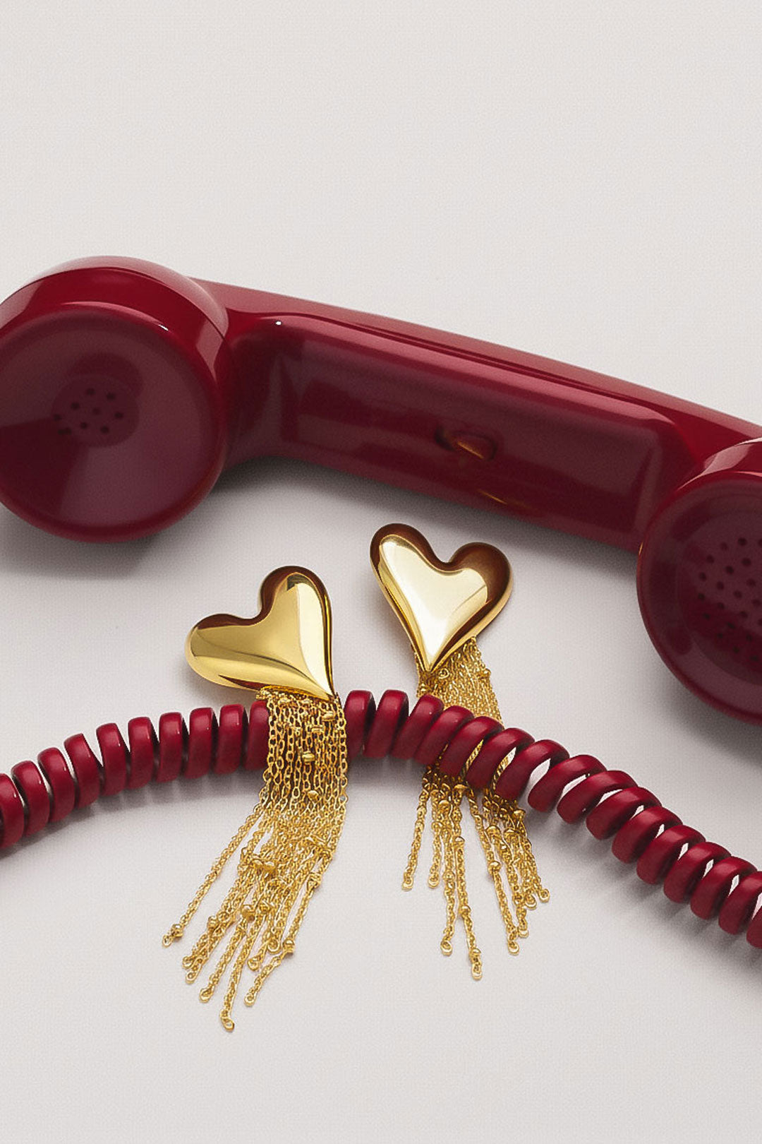 Golden heart dangle earrings placed next to red vintage telephone cord and handset on white surface
