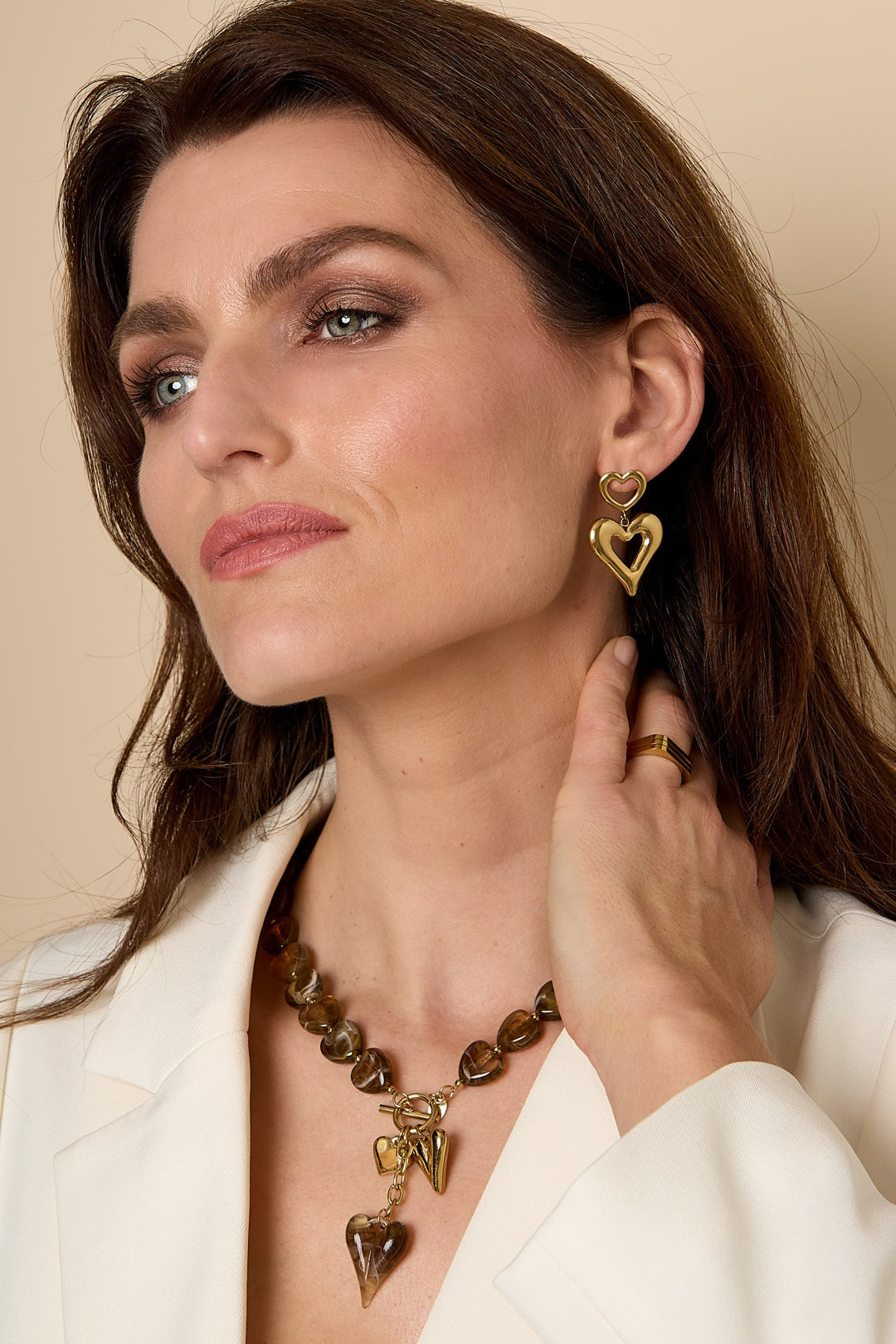 Woman wearing gold double heart drop earrings paired with brown heart necklace and gold ring, dressed in white blazer