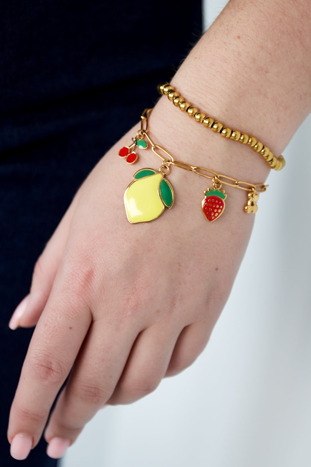 Close-up of a wrist wearing a gold-plated fruit charm bracelet alongside a gold beaded bracelet