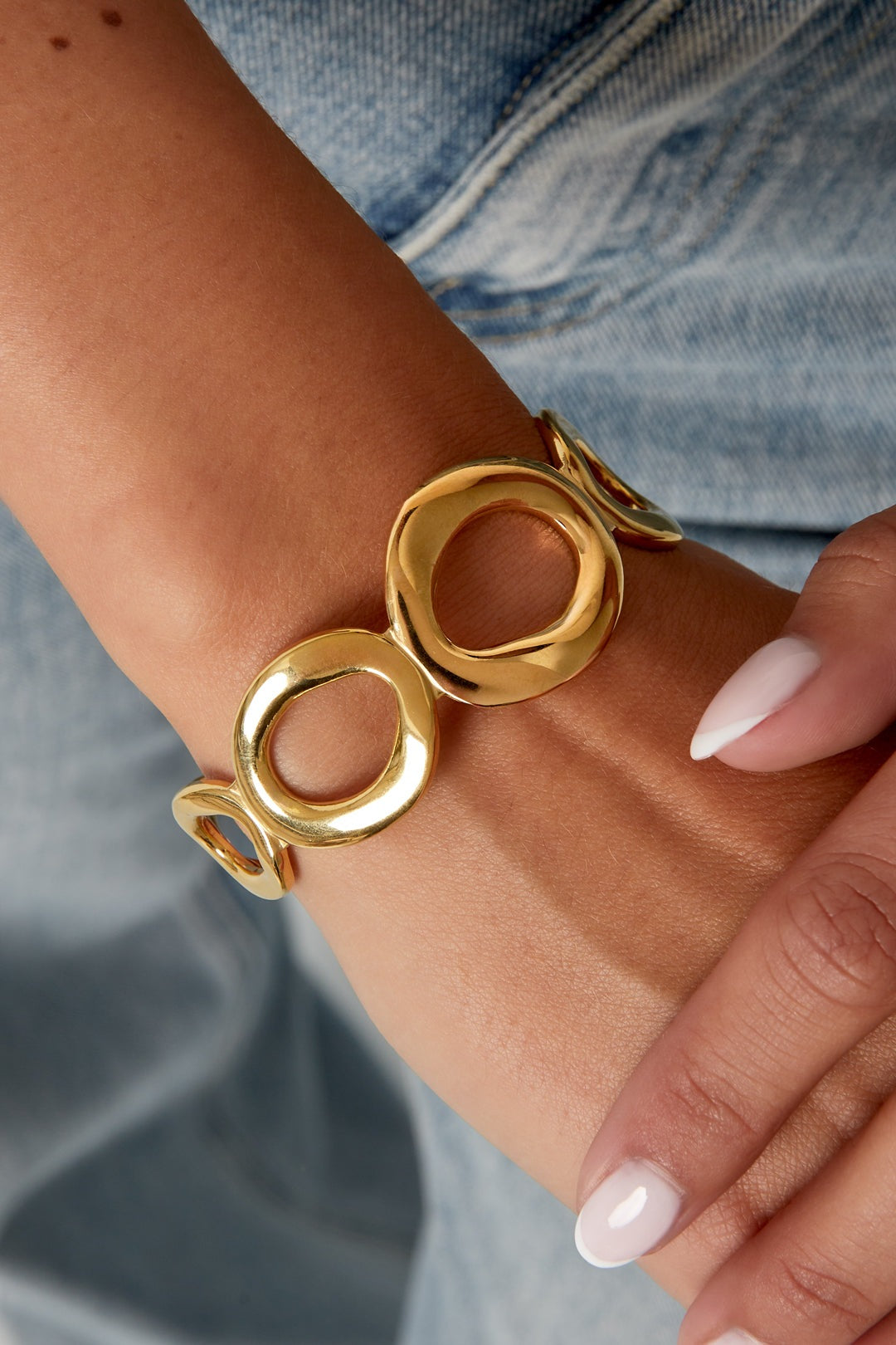 Close-up of a woman's wrist wearing a shiny gold openwork cuff bracelet featuring irregular open oval shapes paired with denim outfit