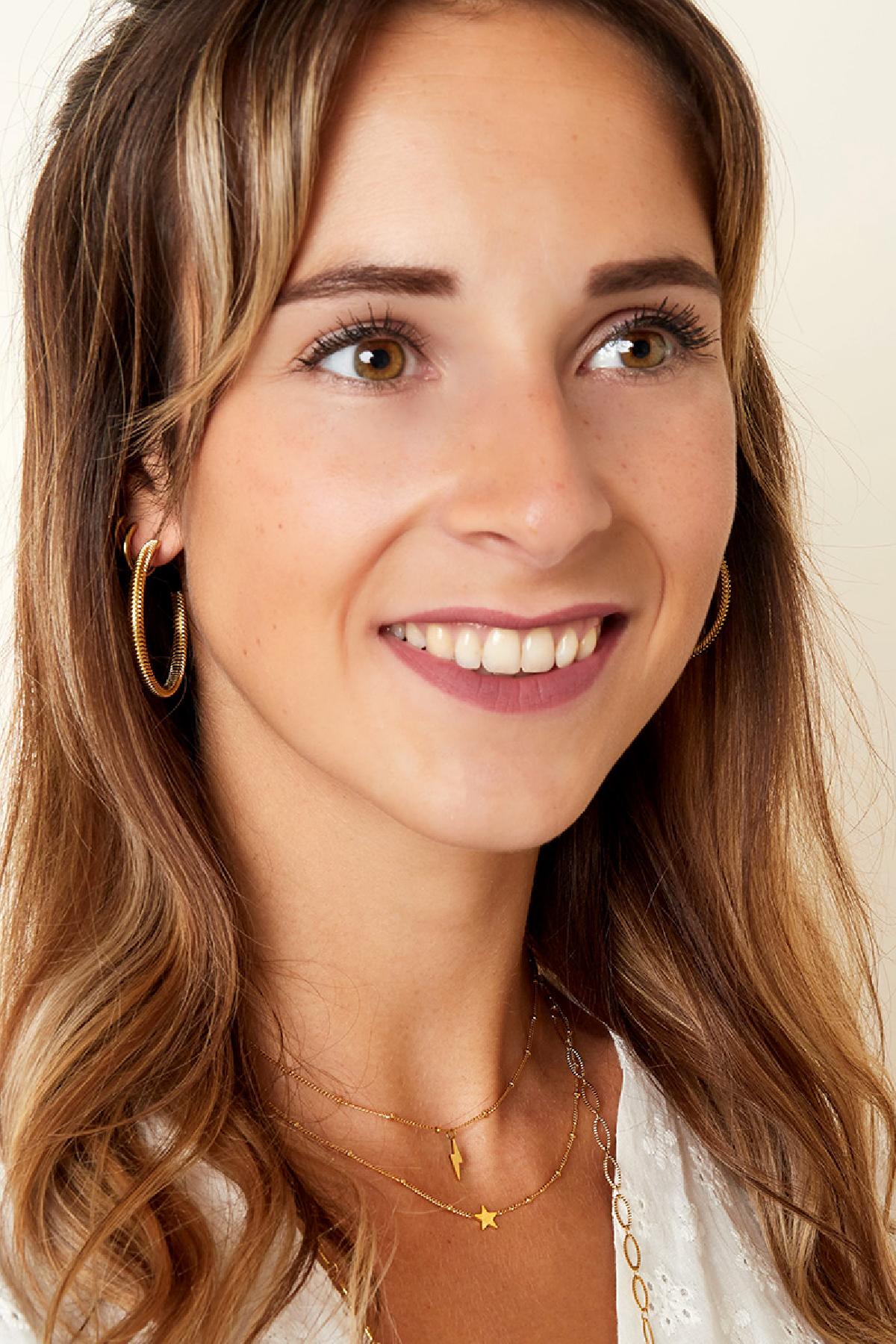 Smiling woman wearing layered gold necklaces with star and lightning bolt pendants, paired with large gold hoop earrings and a white top