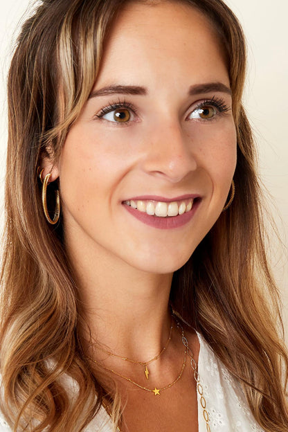 Smiling woman wearing layered gold necklaces with star and lightning bolt pendants, paired with large gold hoop earrings and a white top