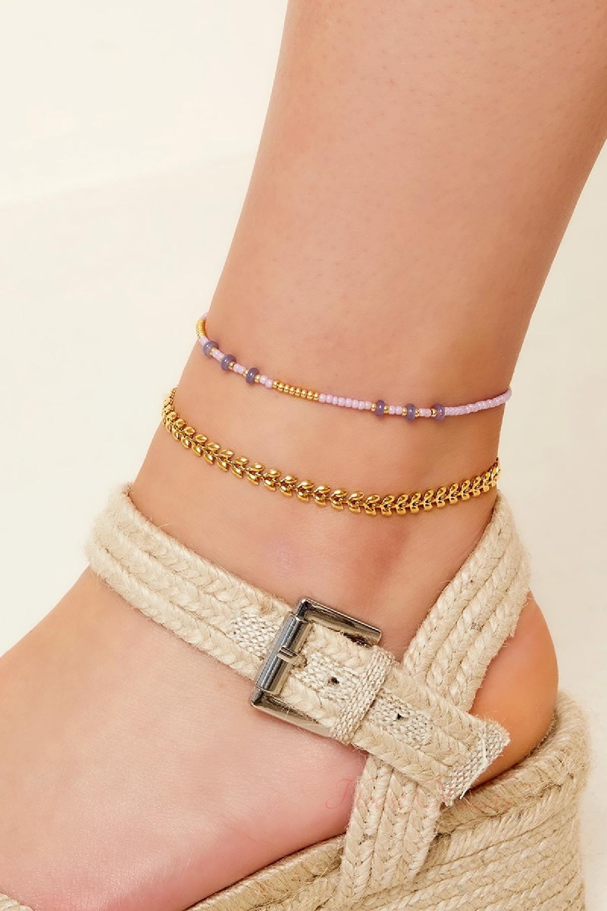 Close-up of a foot wearing two anklets: a gold leaf chain anklet and a delicate beaded lavender and gold anklet, paired with woven wedge sandals