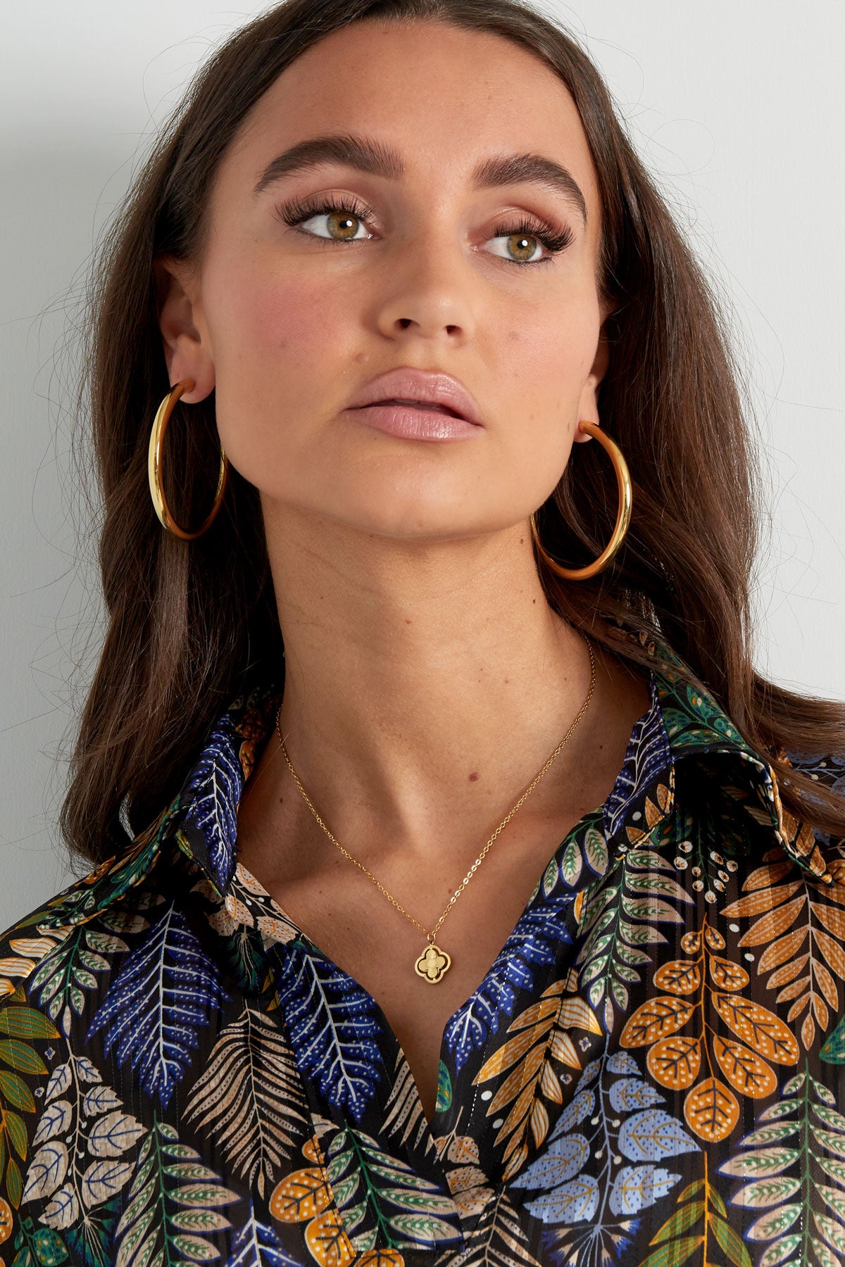 Woman wearing gold clover pendant necklace with blue and orange leaf patterned blouse and large gold hoop earrings