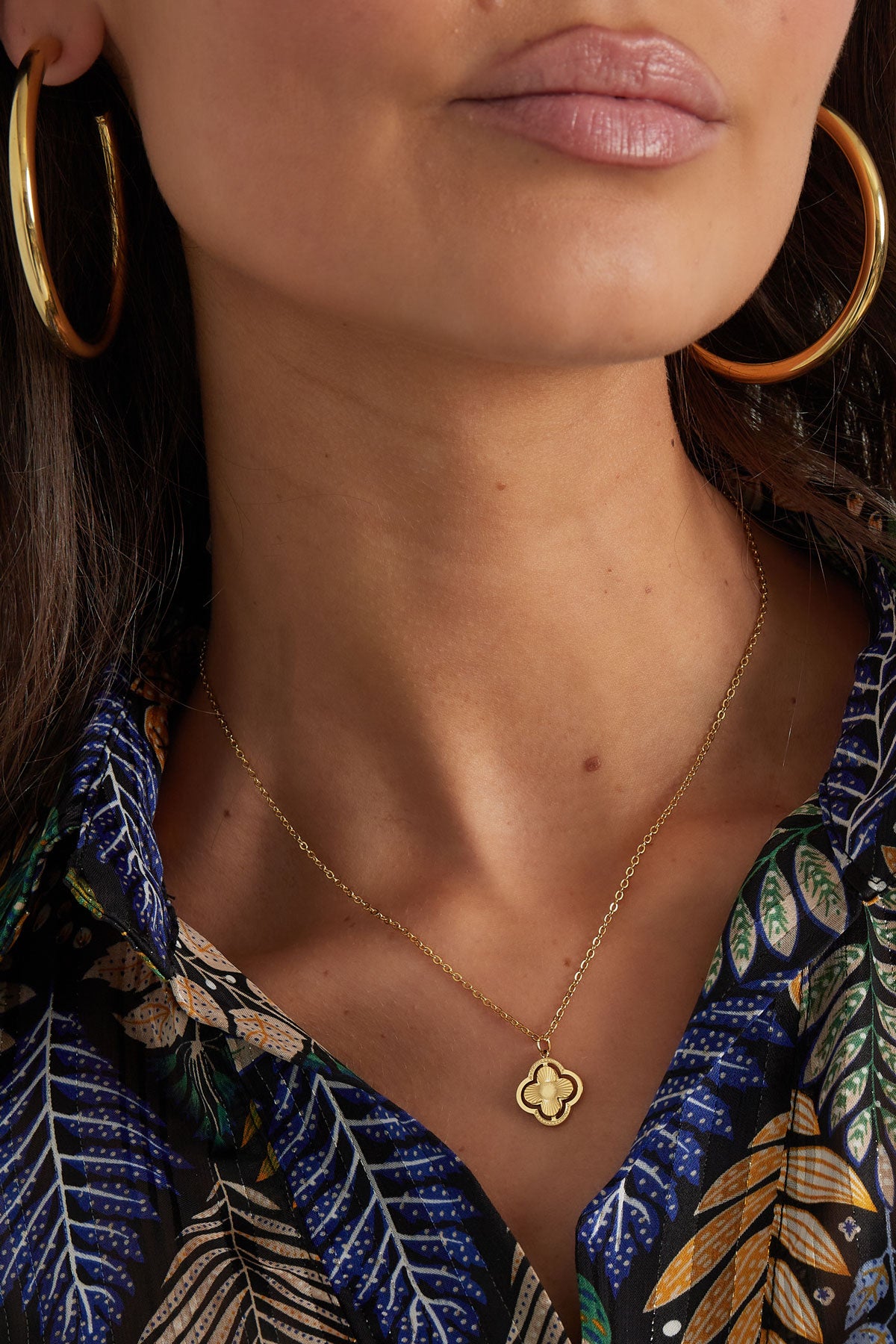 Close-up of woman’s neck with gold clover pendant necklace and large gold hoop earrings, wearing a colourful leaf patterned blouse