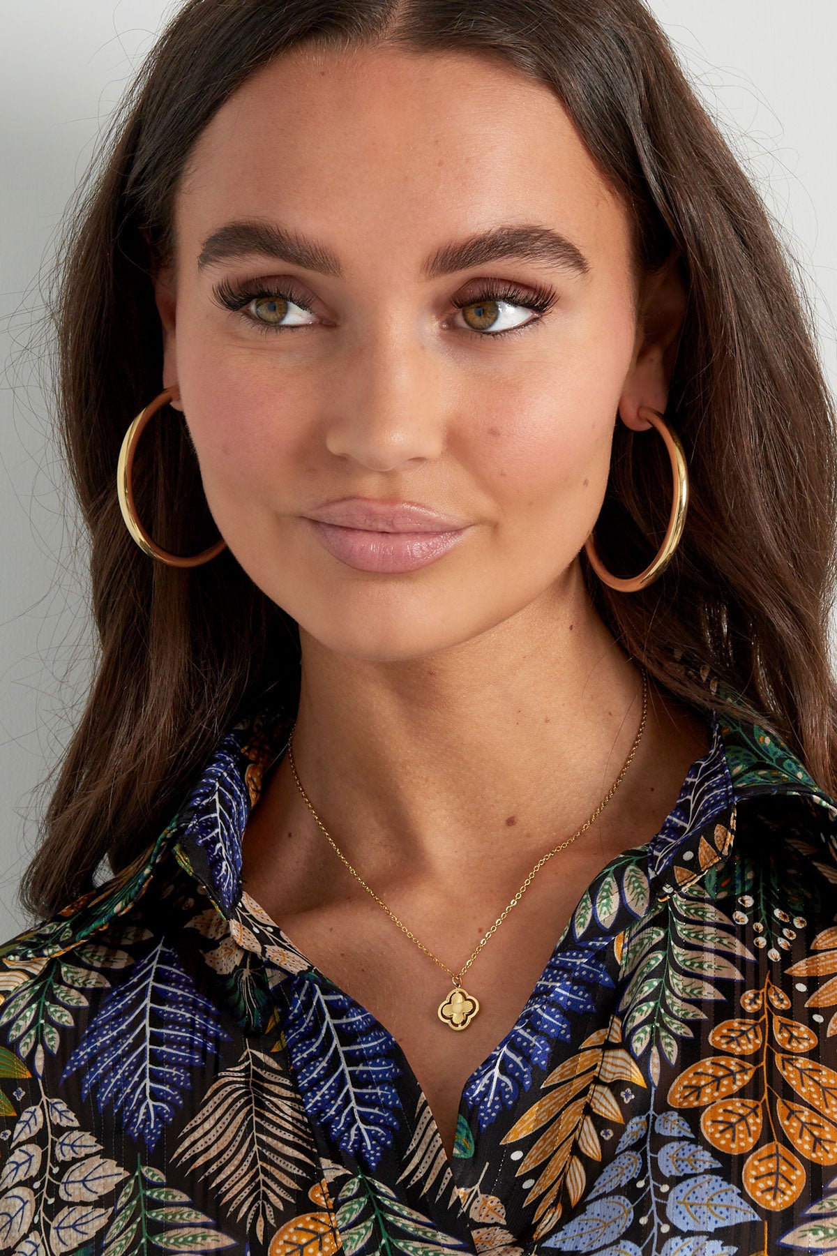 Woman smiling, wearing gold clover pendant necklace and matching large gold hoop earrings with patterned blouse