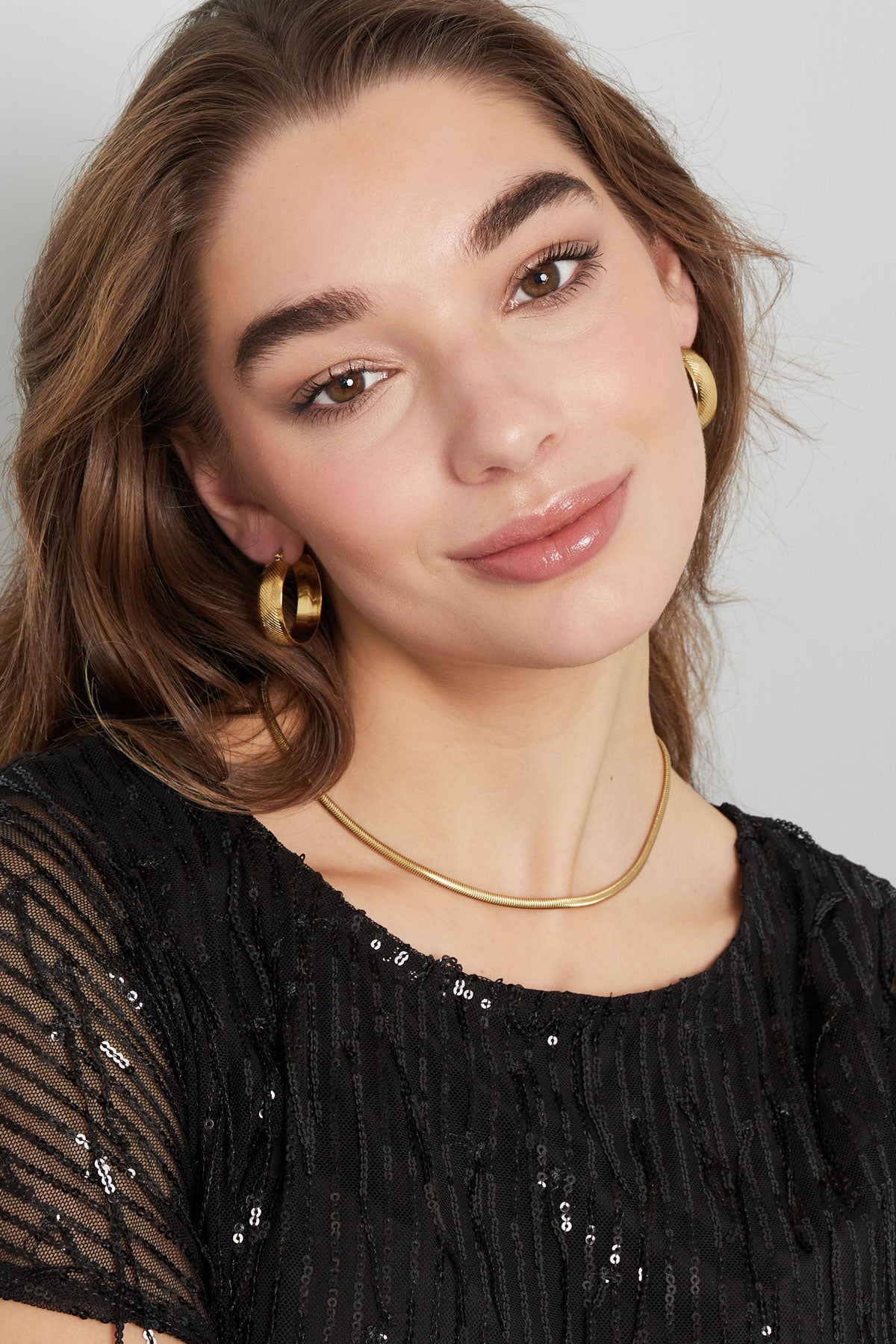 Model wearing gold snake chain necklace paired with hoop earrings and a black sequin top