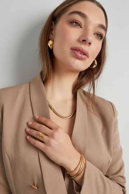Model wearing the gold flat chain necklace paired with matching gold hoop earrings, rings, and bracelets, styled with a beige blazer