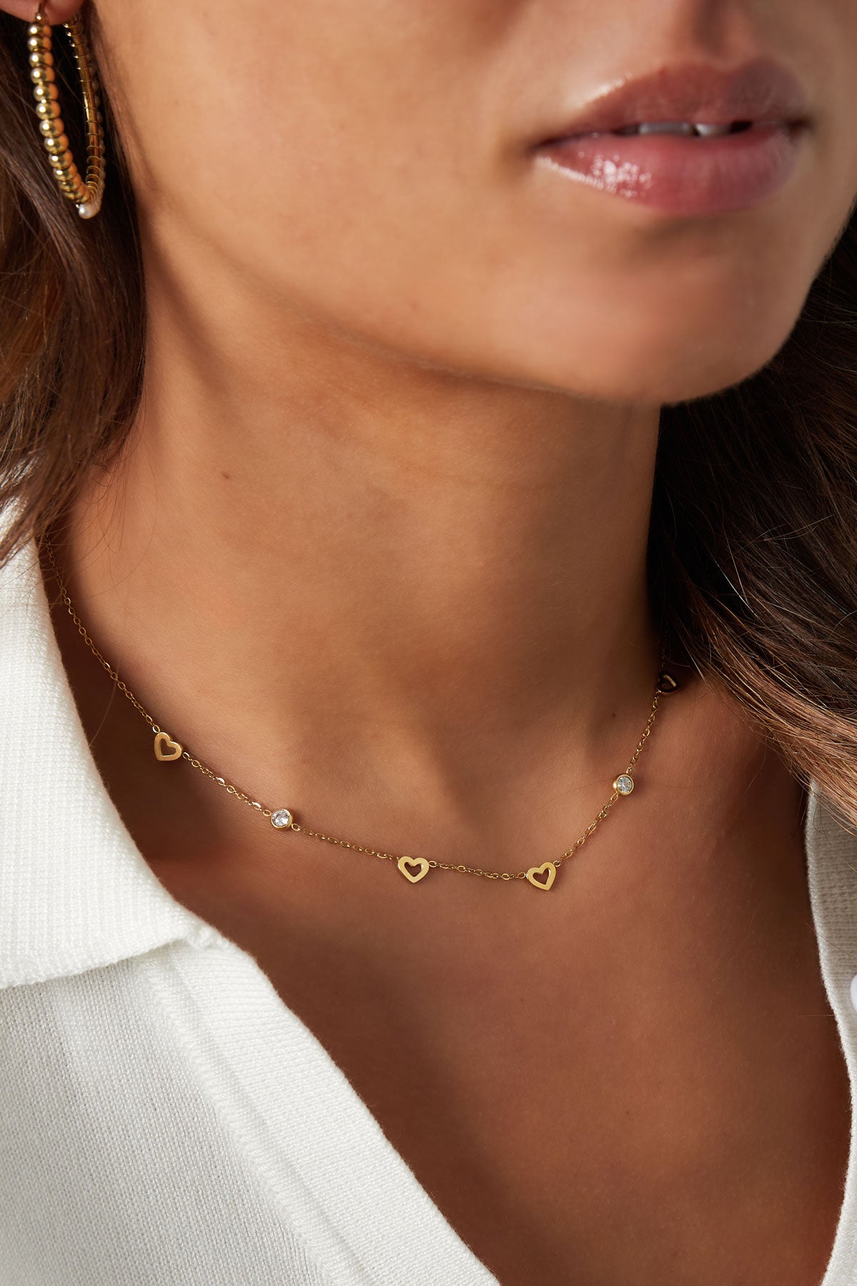 Detailed side view of the gold heart charm necklace worn by a woman in a white outfit, highlighting the delicate design