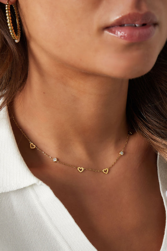 Detailed side view of the gold heart charm necklace worn by a woman in a white outfit, highlighting the delicate design