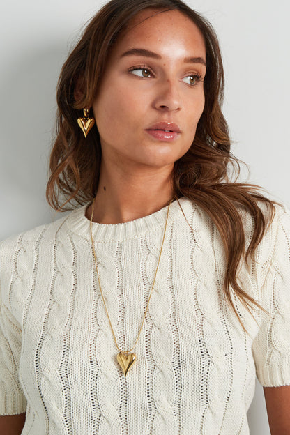 Woman wearing gold chunky heart pendant necklace paired with matching heart earrings and white cable knit sweater