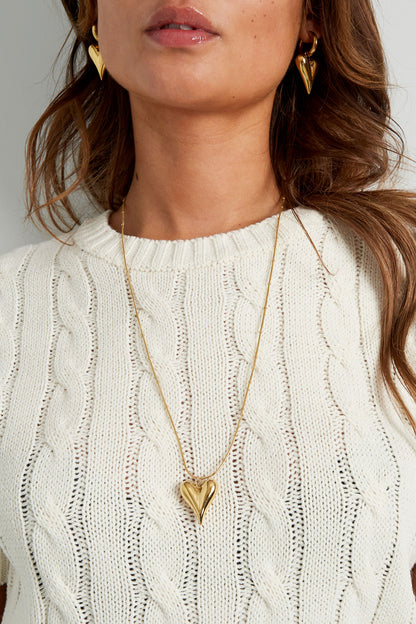 Close-up of woman wearing gold chunky heart pendant necklace and matching earrings, highlighting pendant detail