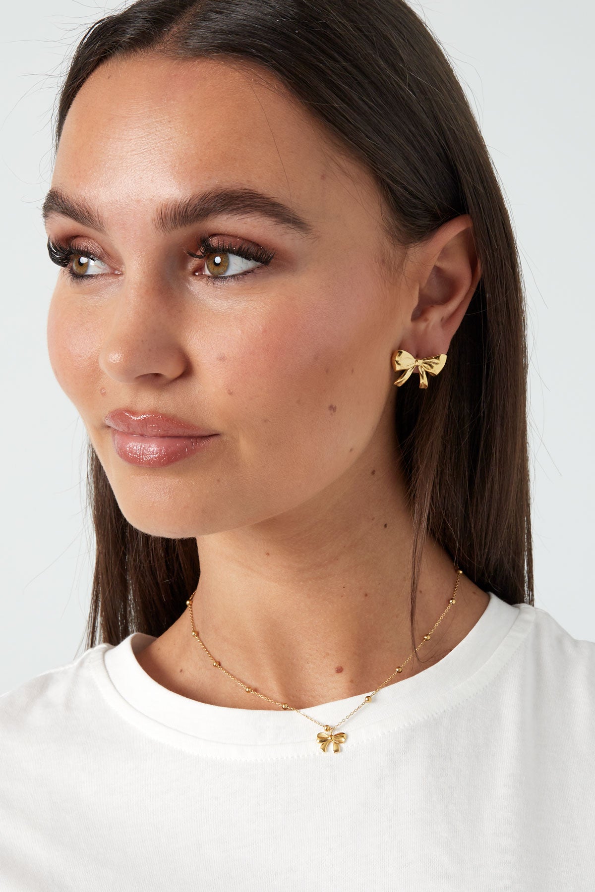 Close-up of a woman wearing gold bow pendant necklace and matching bow-shaped earrings with smooth skin and white top