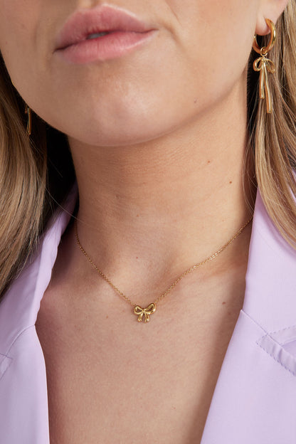Close-up of a woman wearing a minimal gold bow necklace and matching gold earrings with a lilac blazer