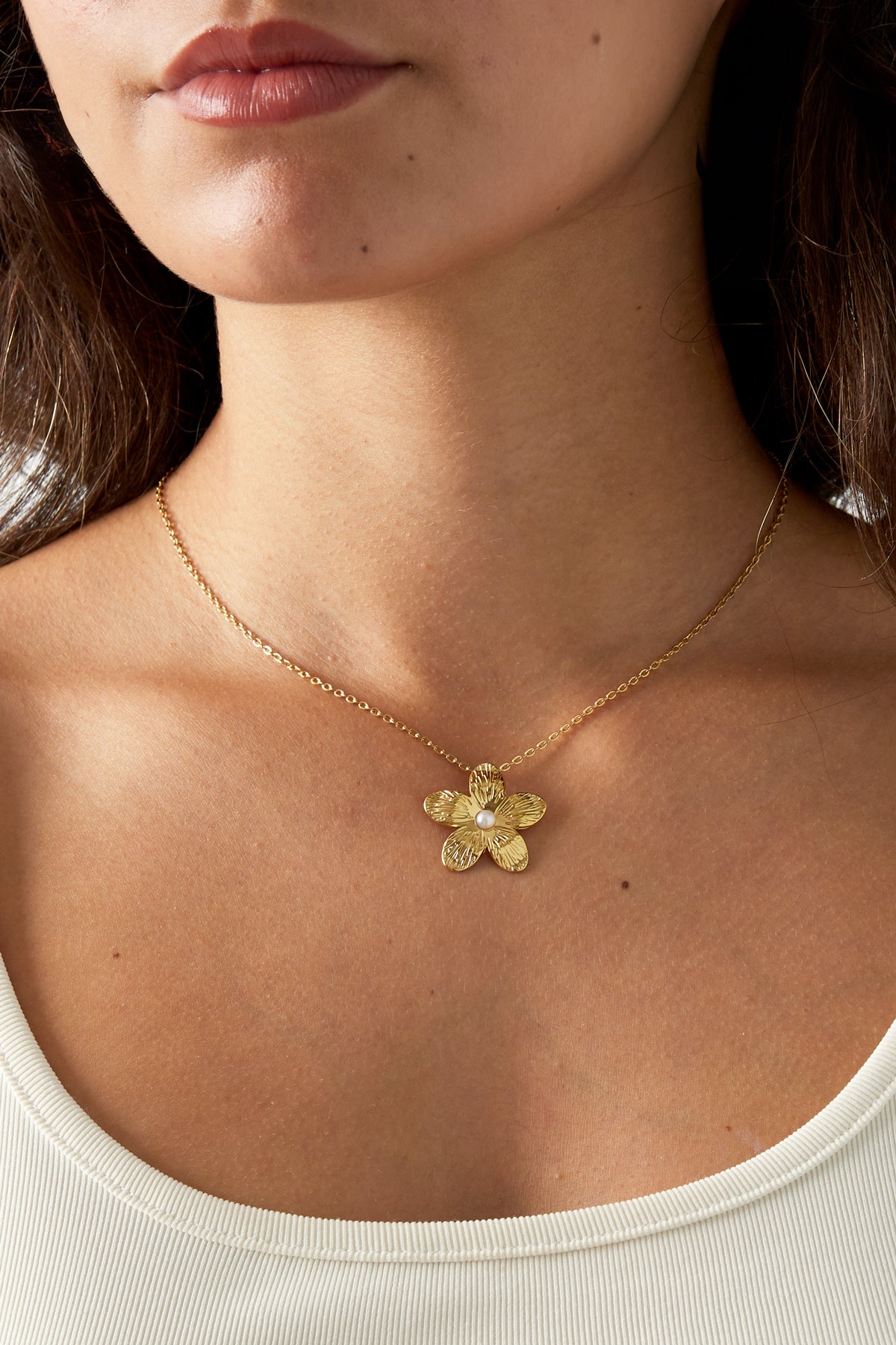 Close-up of a woman wearing a gold flower necklace with pearl detail against bare skin and a white top