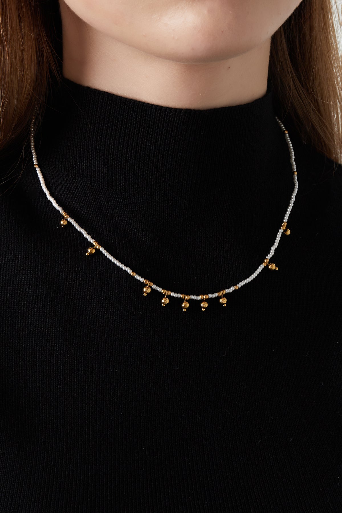 Model wearing delicate white bead necklace with gold bell charms paired with a black top showcasing necklace fit and style