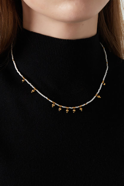 Model wearing delicate white bead necklace with gold bell charms paired with a black top showcasing necklace fit and style