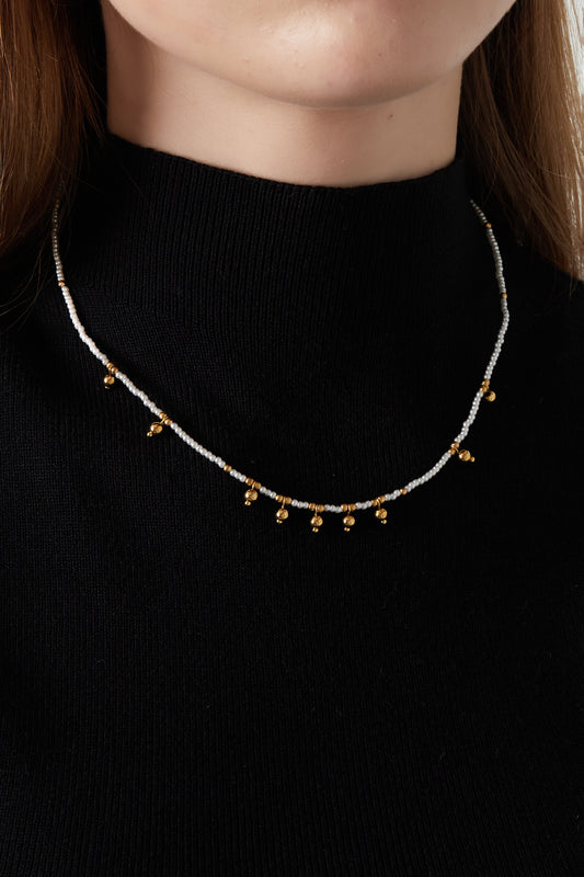 Model wearing delicate white bead necklace with gold bell charms paired with a black top showcasing necklace fit and style