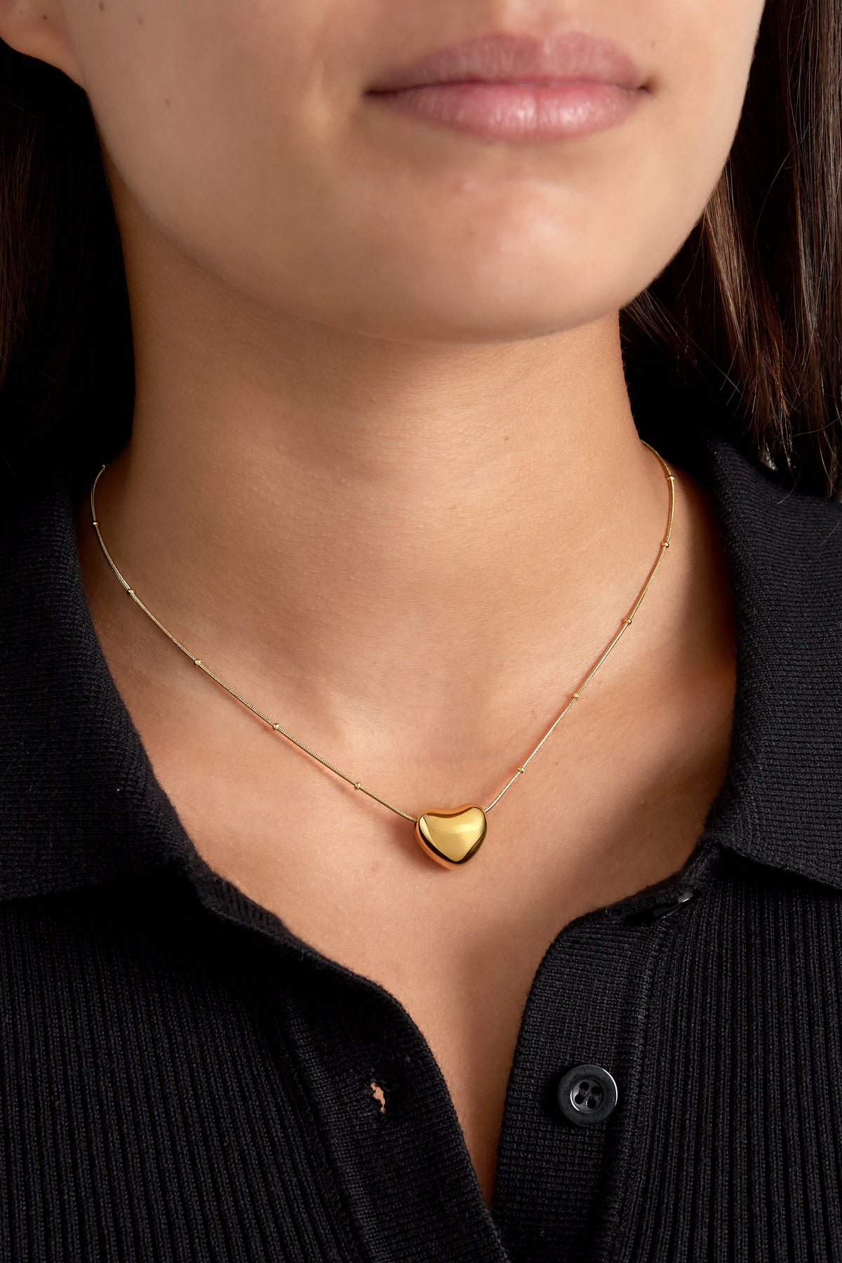 Model wearing a gold heart pendant necklace over a black top, highlighting the soft polished pendant at clavicle level