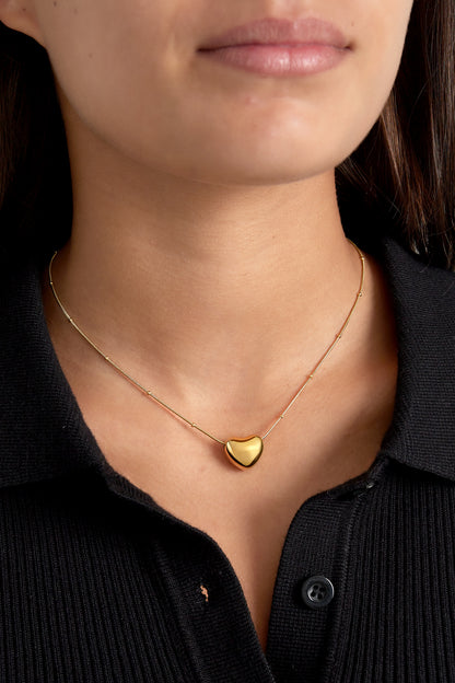 Model wearing a gold heart pendant necklace over a black top, highlighting the soft polished pendant at clavicle level