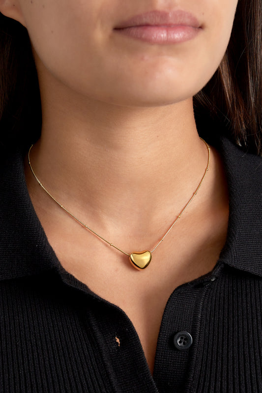 Model wearing a gold heart pendant necklace over a black top, highlighting the soft polished pendant at clavicle level