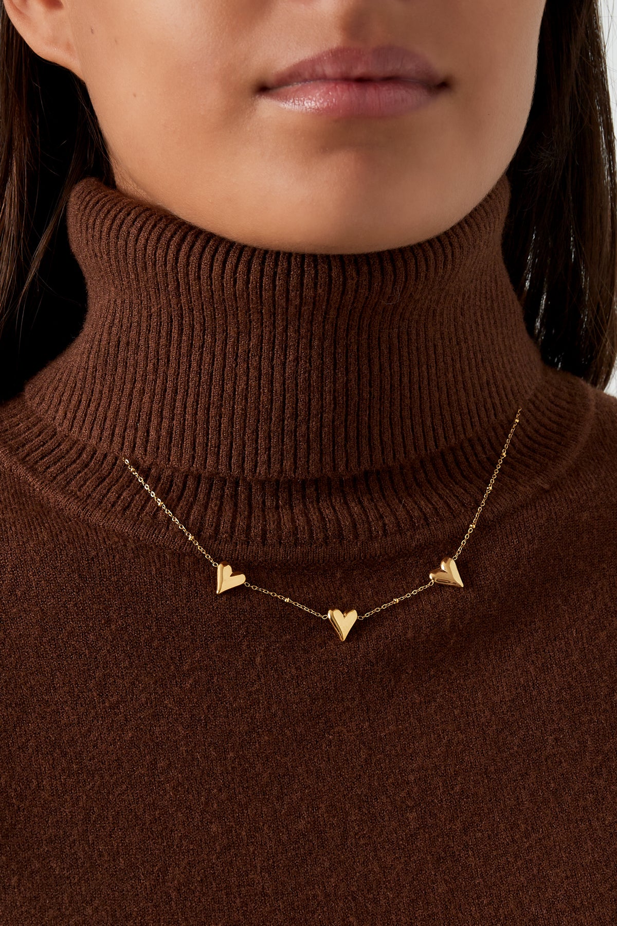 Close-up image of a woman wearing the triple heart gold necklace over a brown ribbed turtleneck sweater, showcasing the necklace's elegant fit