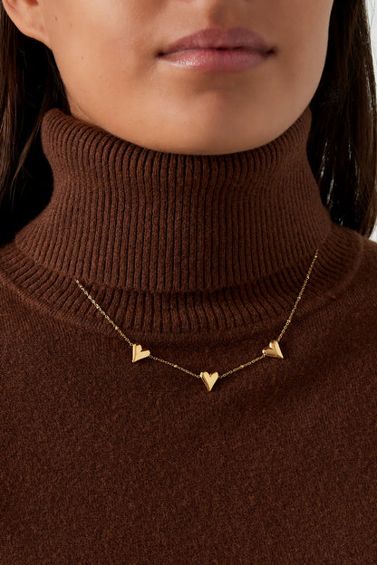 Close-up image of a woman wearing the triple heart gold necklace over a brown ribbed turtleneck sweater, showcasing the necklace's elegant fit