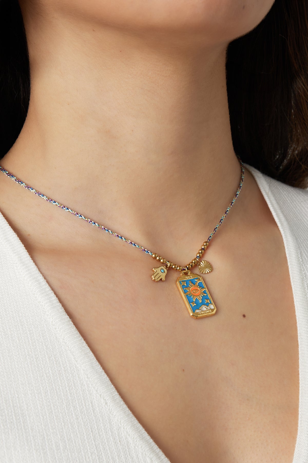 Model wearing braided necklace with gold sun, heart, and Hamsa hand charms against white top
