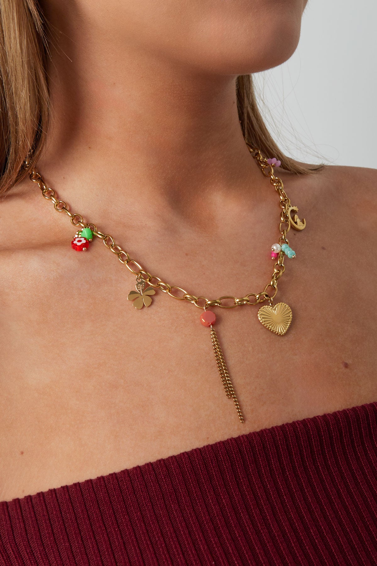 Model wearing gold charm chain necklace featuring heart pendant, moon and clover charms with colourful bead details, styled with burgundy top