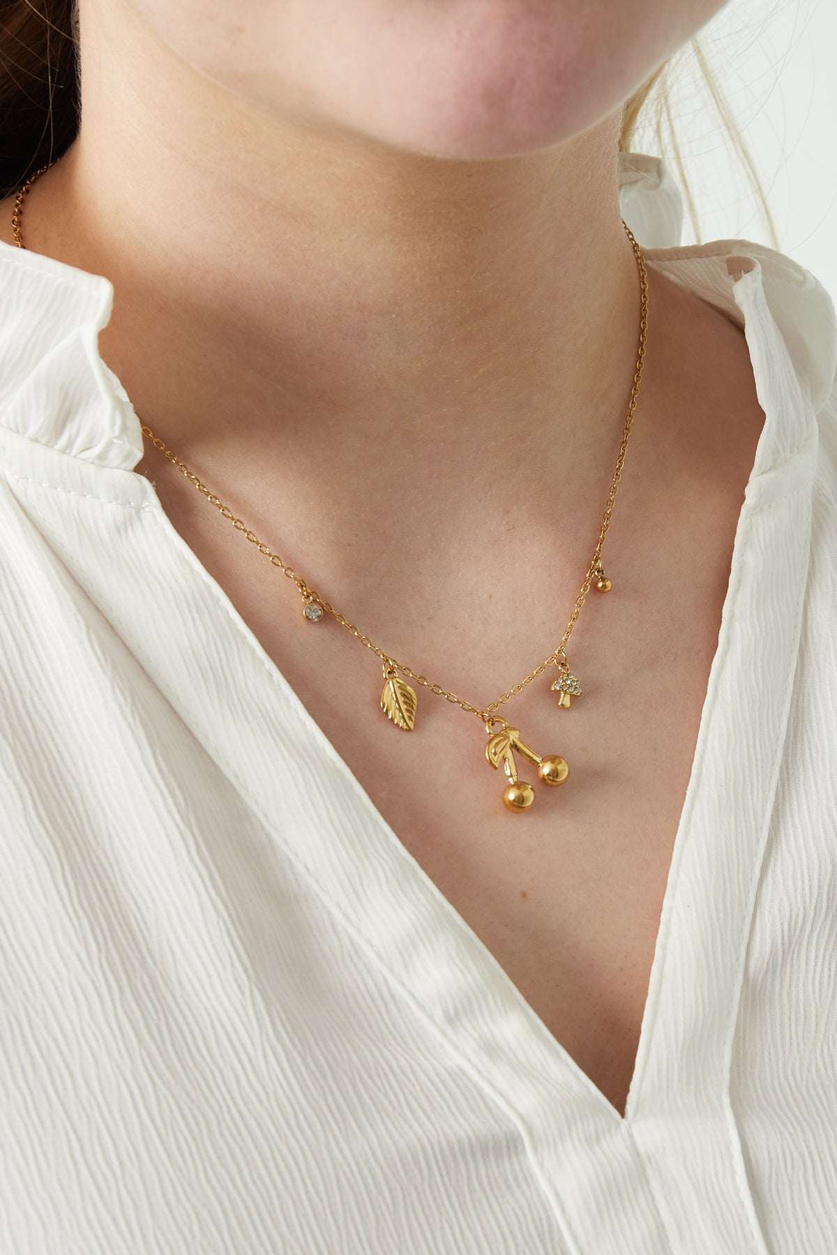 Close-up of a woman wearing a gold charm necklace with cherry and nature-inspired pendants paired with a white textured blouse
