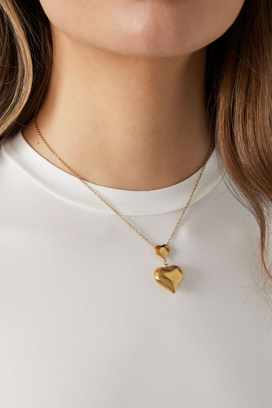 Close-up of a woman wearing the gold double heart pendant necklace over a white top, showing the pendant resting near the collarbone