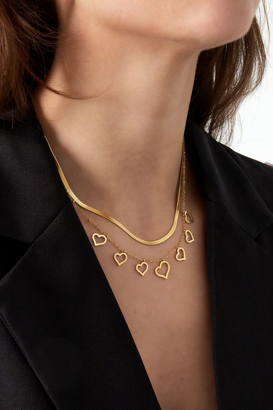 Model wearing the gold layered heart necklace pairing a smooth snake chain and a charm chain with seven hearts on neckline with a black outfit