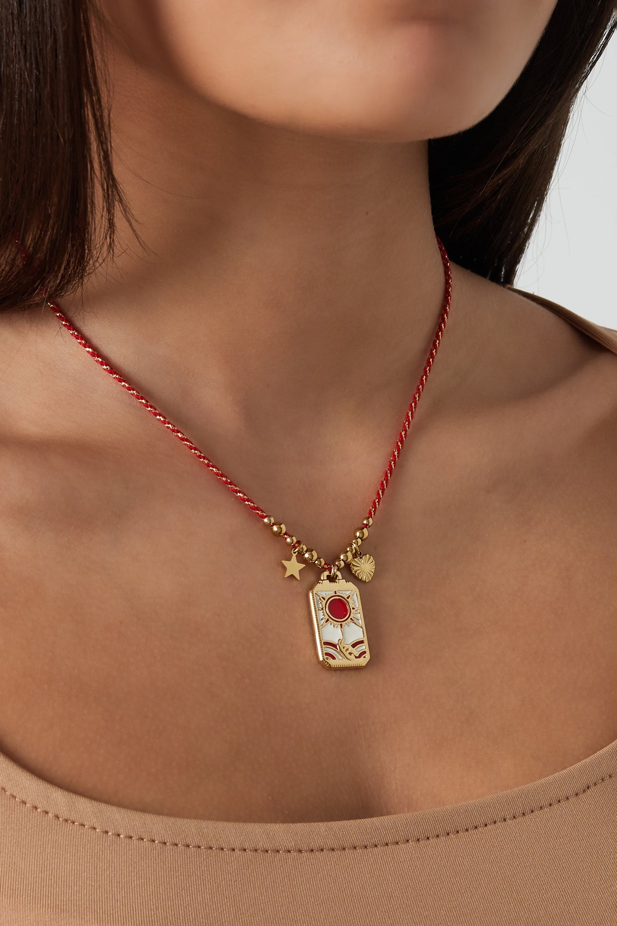 Model wearing solar charm necklace with red braided cord and gold-tone sun, hand, star and heart charms, styled with beige top