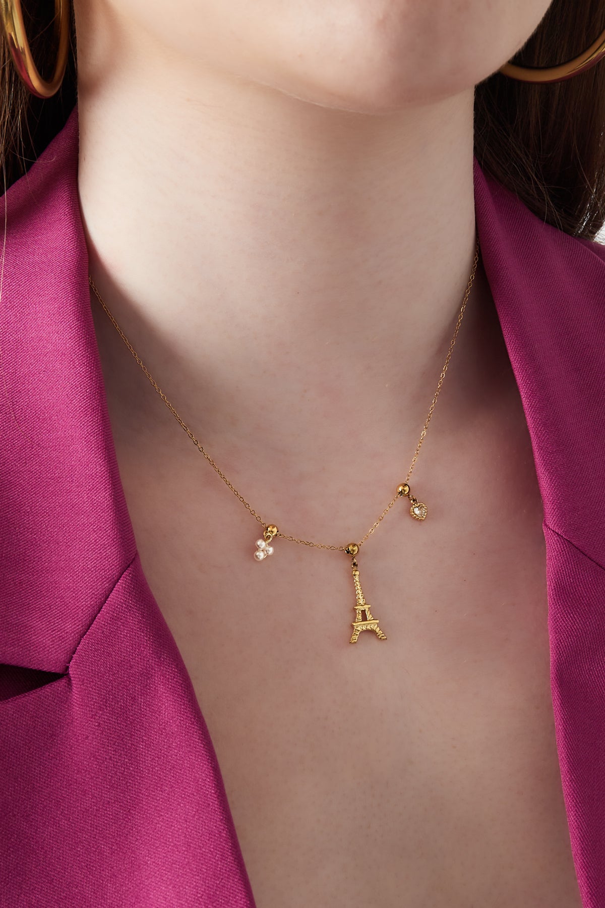 Close-up of woman wearing gold Eiffel Tower charm necklace with pearl cluster and heart charms against purple blazer