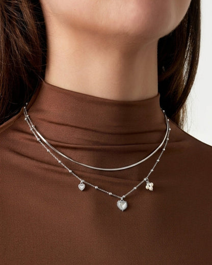Silver coquette layered necklace with heart and pearl charms, worn against a brown top.
