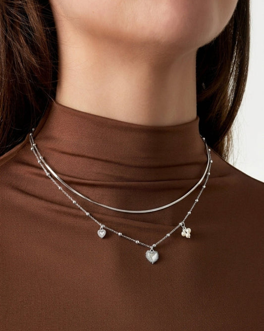 Silver coquette layered necklace with heart and pearl charms, worn against a brown top.