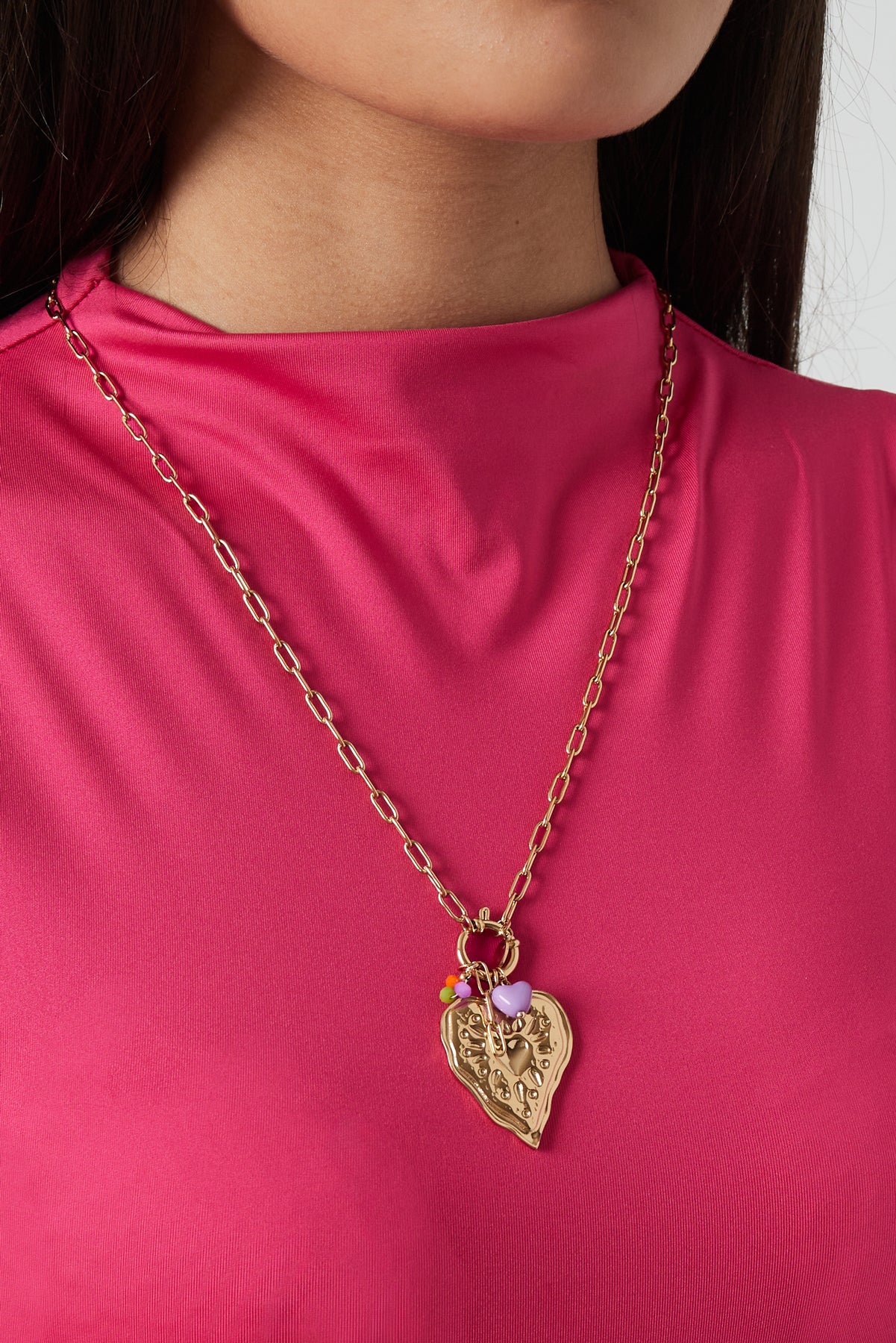 Close-up of model wearing gold heart pendant necklace with purple and colourful charms paired with bright pink top