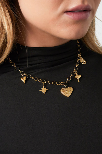 Close-up of a woman wearing a gold-tone charm necklace with heart, star, ballerina, and tree charms over a black high-neck top