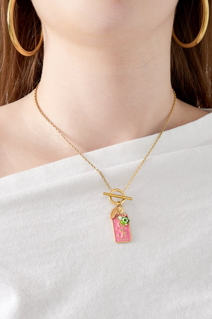 Close-up of the pendant necklace worn on a woman dressed in white, showcasing the pink floral tag and gold toggle clasp
