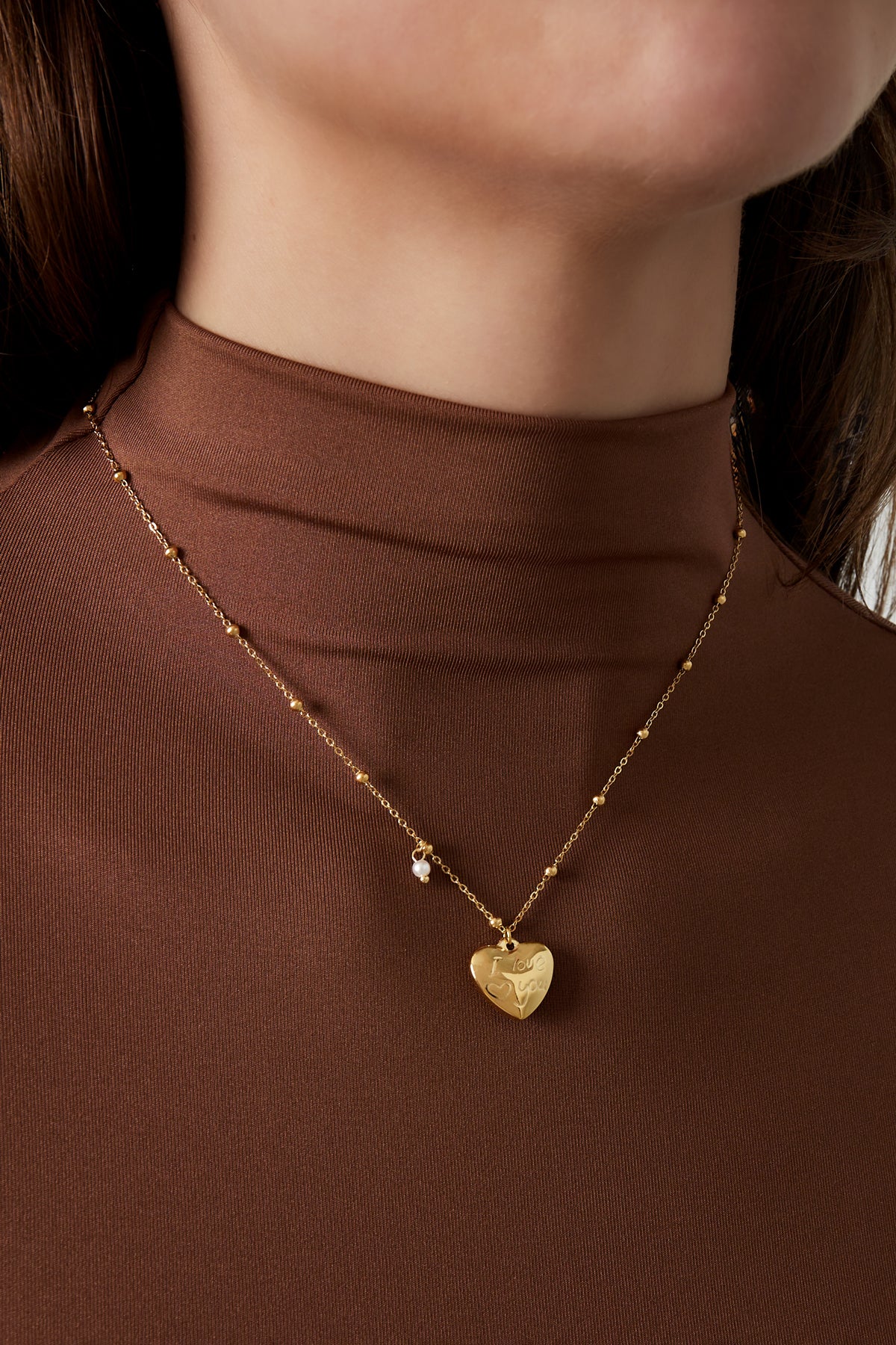 Close-up of woman wearing gold heart charm pendant necklace with brown top