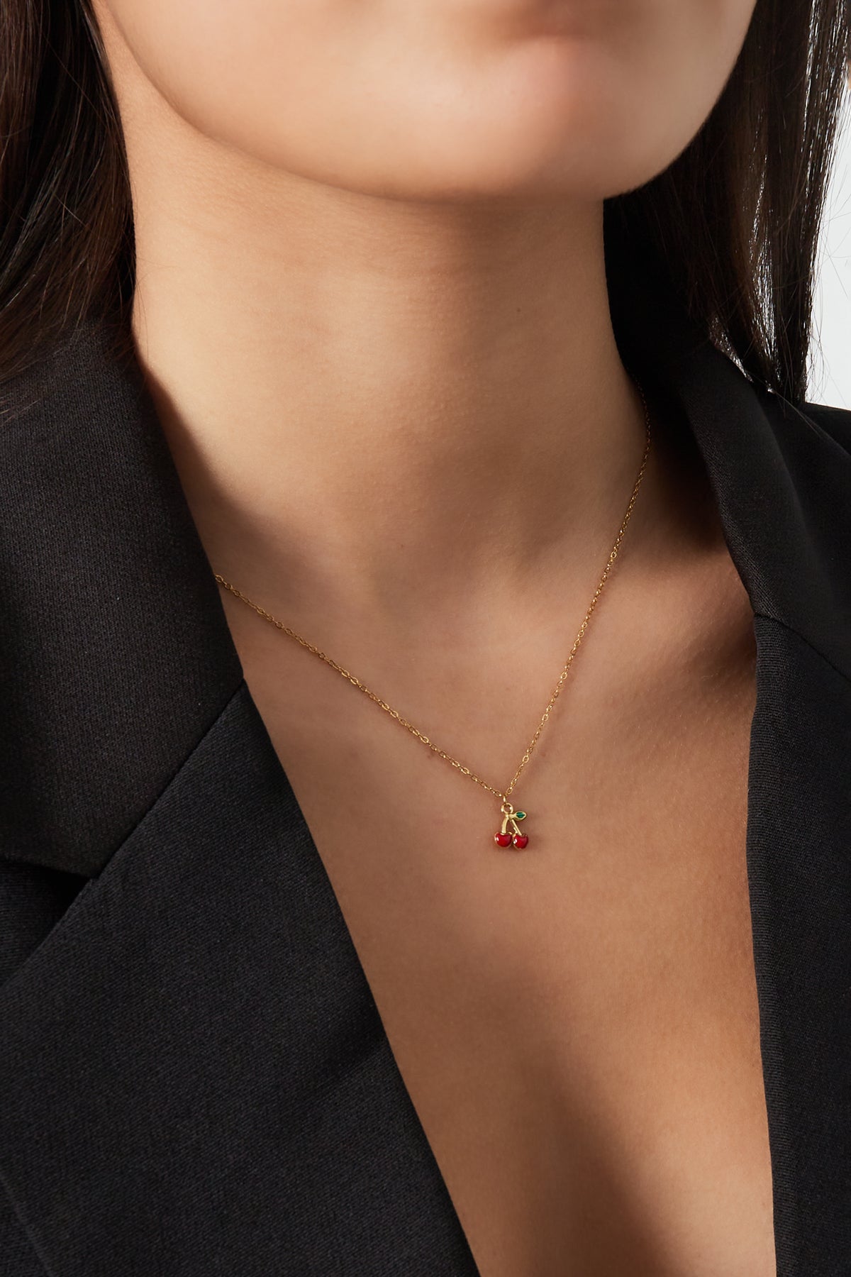 Close-up of woman wearing a gold-coloured chain necklace featuring a red enamel cherry pendant, styled with a black blazer