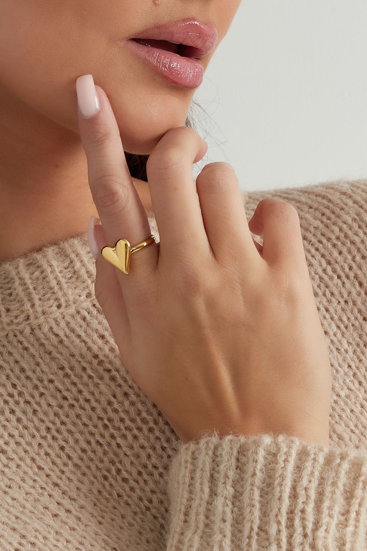 Woman wearing a gold heart ring on her finger, touching her face, dressed in a soft beige knit sweater