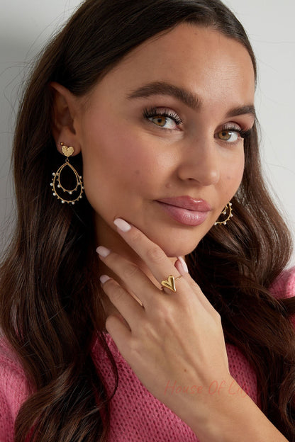 Smiling woman wearing gold heart ring on finger and matching gold earrings with pearls against white background