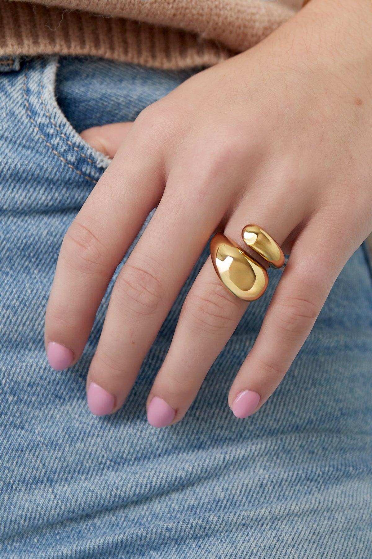 Hand wearing a shiny gold sculptural ring against blue denim jeans and beige knitwear