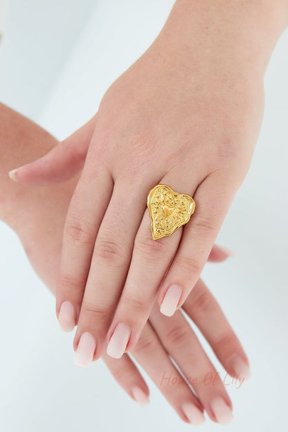 Woman's hand wearing gold wrinkled heart statement ring with manicured nails against light background