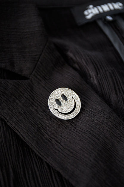 Silver-tone smiley face brooch with clear crystals pinned to a black textured jacket