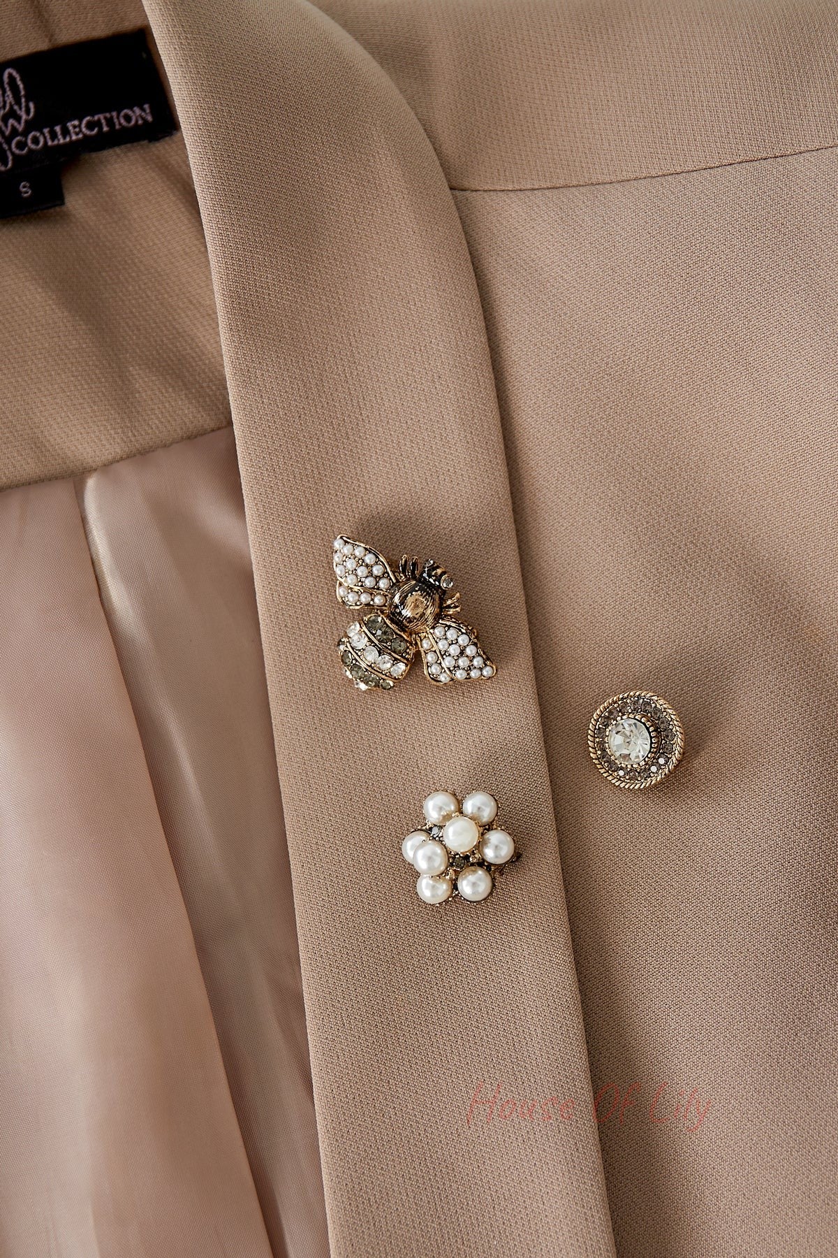 Three brooches pinned on a beige blazer lapel: a pearl and rhinestone bee, a circular crystal brooch, and a pearl cluster flower emphasizing their boutique style