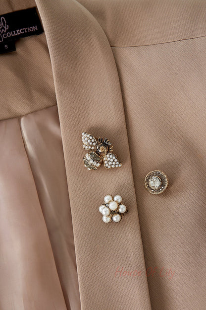 Three brooches pinned on a beige blazer lapel: a pearl and rhinestone bee, a circular crystal brooch, and a pearl cluster flower emphasizing their boutique style