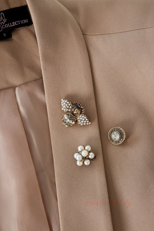 Three brooches pinned on a beige blazer lapel: a pearl and rhinestone bee, a circular crystal brooch, and a pearl cluster flower emphasizing their boutique style